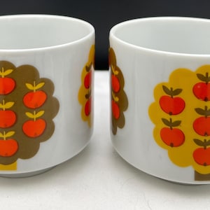 May include: Two white ceramic mugs with a rounded shape. Each mug features a stylized tree design with orange apples and brown or yellow foliage. The mugs are suitable for coffee, tea, or other beverages.