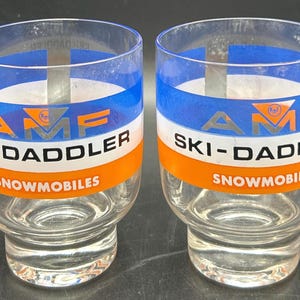 May include: Two clear glass tumblers with a blue, white, and orange band design. The text "AMF SKI-DADDLER SNOWMOBILES" is printed on the front of each glass. The glasses have a rounded base.