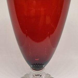 Puede incluir: Una copa de cristal rojo rubí con una base decorativa transparente. La copa tiene un cuenco ancho y cónico y una base circular transparente con pequeños adornos redondos. El cristal tiene una superficie lisa y reflectante.