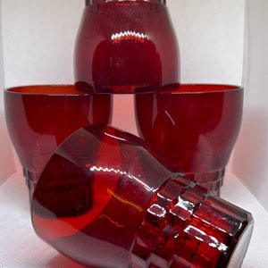 Puede incluir: Juego de cinco vasos de color rojo rubí. Cuatro vasos están en posición vertical y uno está de lado. Los vasos tienen un diseño geométrico en la base y un cuenco redondeado. Los vasos son de material transparente.