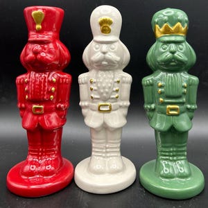 May include: Three ceramic nutcracker figurines in red, white, and green. Each figurine is in the shape of a soldier with a dog-like face, wearing a hat and uniform with gold accents. The figurines are standing on a round base.