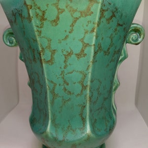 May include: A turquoise ceramic vase with a geometric design and brown speckled accents. The vase has a flared top, a rounded base, and decorative handles. The vase is approximately 25 cm tall.