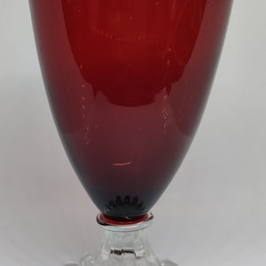 May include: A deep red wine glass with a clear, spherical base. The glass has a wide, conical bowl and a clear stem with a ring of clear, round beads. The glass is likely made of glass.
