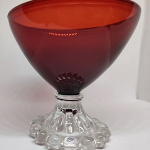 May include: A ruby red glass goblet with a wide, conical bowl and a clear, decorative base. The bowl has a smooth surface, while the base features a ring of clear glass spheres. The goblet is likely used for serving beverages.