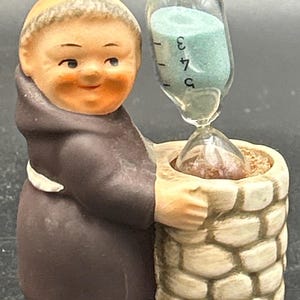 May include: A ceramic figurine of a monk wearing a brown hooded robe, standing beside a stone-textured well. The monk holds a clear glass hourglass containing light blue sand, with the numbers 3, 4, and 5 printed on the glass surface.