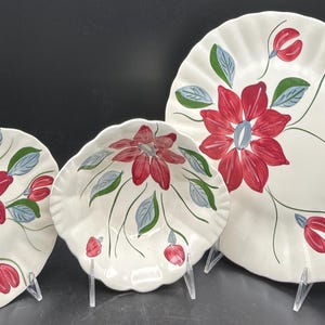 May include: Set of three ceramic plates with a floral design. The plates are white with a red flower, blue center, and green leaves. The plates are different sizes and shapes, with a scalloped edge.