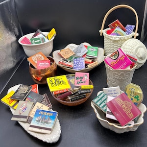 Puede incluir: Una colección de cajas de cerillas antiguas en varios recipientes. Las cajas de cerillas presentan diseños y textos coloridos, incluyendo "Ruby Falls", "Harold's" y "Excalibur". Los recipientes incluyen una cesta blanca, una taza de cobre y cuencos decorativos.