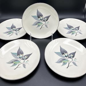 May include: A set of five white ceramic plates with a silver rim. Each plate features a central design of stylized leaves in shades of gray, teal, and green, with small white flowers. The plates are arranged on a dark surface.