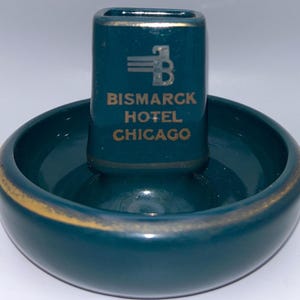 Puede incluir: Un cenicero de cerámica turquesa con un portacigarrillos a juego. El portacigarrillos tiene las palabras "Bismarck Hotel Chicago" en letras doradas. El cenicero tiene un borde dorado.