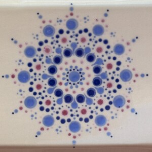 May include: A rectangular, off-white ceramic tray featuring a mandala design. The mandala is composed of blue and red dots of varying sizes, arranged in a circular pattern. The tray has a light brown border.