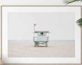 May include: A framed horizontal wall art print featuring a light blue lifeguard tower standing on a sandy beach. The tower has a small American flag attached to a pole on the roof and the text "M 6" visible on the front structure above the window.