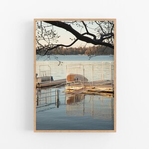 May include: A wooden framed photograph of a lake with a dock and a tree branch extending over the water. The dock is made of wood and metal and has a small boat on it. The water is calm and reflects the sky and trees.