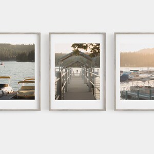 May include: Set of three framed landscape prints featuring serene lake scenes. The prints depict boats docked at a pier, a wooden walkway, and a view of the lake with a boat. The frames are a light wood color.