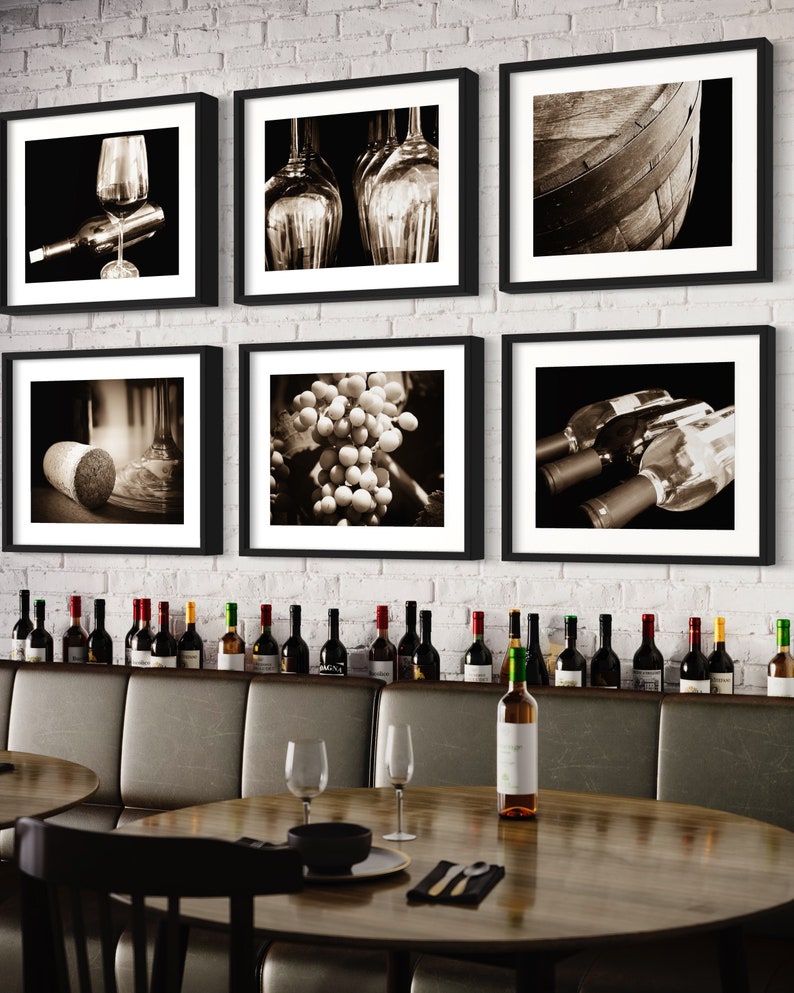 Op de afbeelding: Een set van zes ingelijste zwart-witfoto's die verschillende wijn-gerelateerde afbeeldingen tonen, waaronder druiven, wijnflessen, glazen en een houten vat. De foto's worden weergegeven op een witte bakstenen muur boven een houten tafel met wijnflessen en glazen.