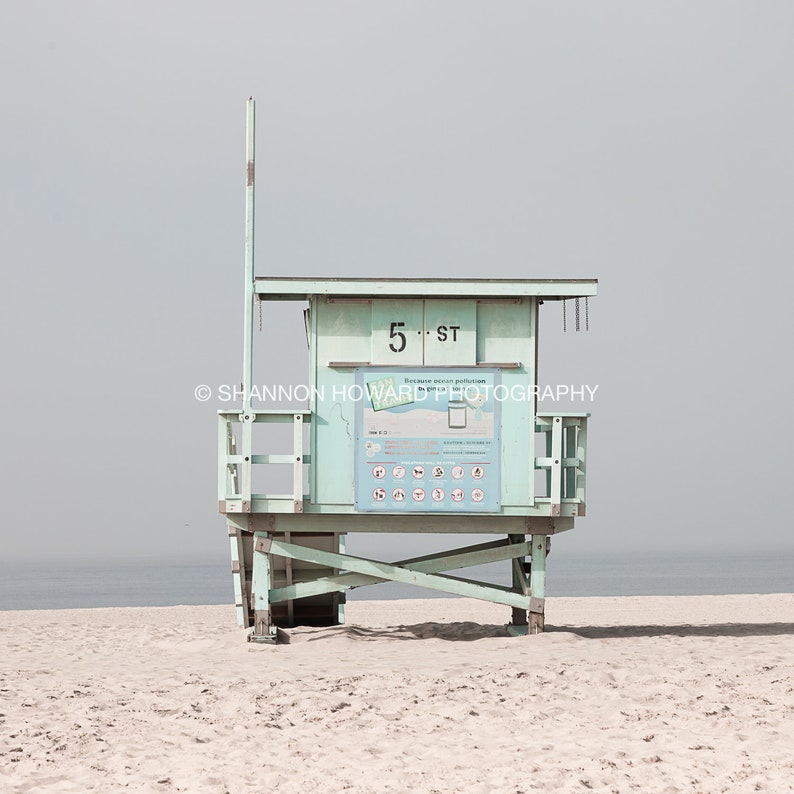 May include: A light blue lifeguard stand with a white sign that reads "5th St. Ocean Safety Patrol" on a sandy beach.