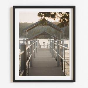 May include: A wooden dock with a covered walkway extends over a calm lake. The dock is lined with railings and has a person standing at the end of the walkway.