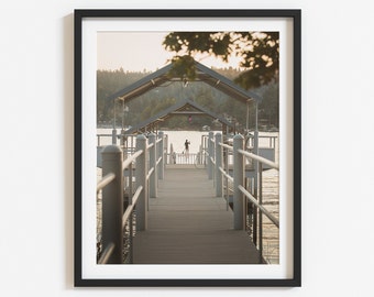May include: A wooden dock with a covered walkway extends over a calm lake. The dock is lined with railings and has a person standing at the end of the walkway.