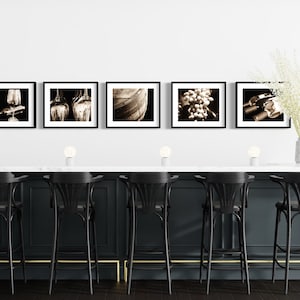 Op de afbeelding: Een zwart-wit fotocollage van wijn gerelateerde beelden, waaronder druiven, wijnflessen, glazen en een vat. De collage wordt weergegeven op een witte muur boven een zwarte bar met gouden accenten. Er zijn zes zwarte barstoelen met gouden accenten.