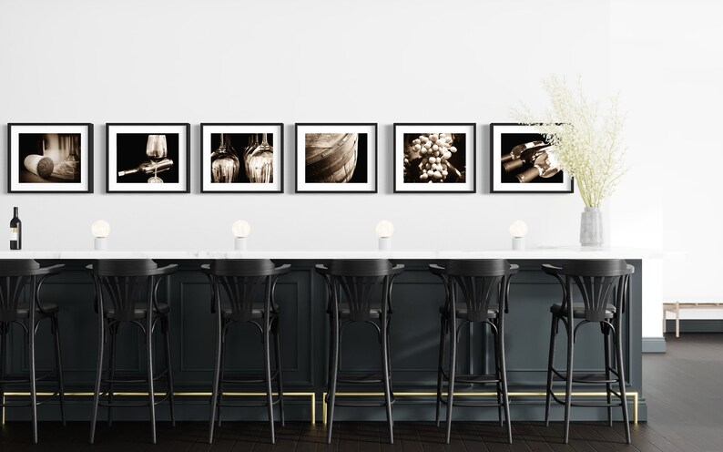 Op de afbeelding: Een zwart-wit fotocollage van wijn gerelateerde beelden, waaronder druiven, wijnflessen, glazen en een vat. De collage wordt weergegeven op een witte muur boven een zwarte bar met gouden accenten. Er zijn zes zwarte barstoelen met gouden accenten.