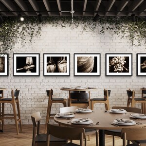 Op de afbeelding: Een restaurantinterieur met een bakstenen muur en een rij ingelijste zwart-witfoto's van wijn gerelateerde items, waaronder druiven, flessen en vaten. De foto's hangen boven een houten tafel met stoelen.
