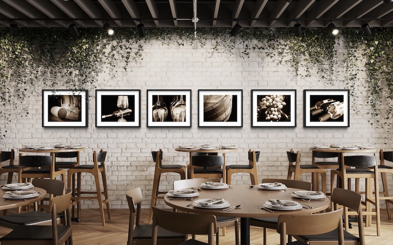 Op de afbeelding: Een restaurantinterieur met een bakstenen muur en een rij ingelijste zwart-witfoto's van wijn gerelateerde items, waaronder druiven, flessen en vaten. De foto's hangen boven een houten tafel met stoelen.