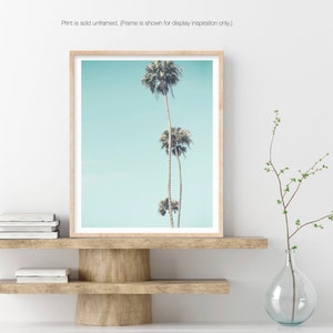May include: A framed print of three palm trees against a light blue sky. The print is in a light wood frame and is displayed on a wooden shelf.