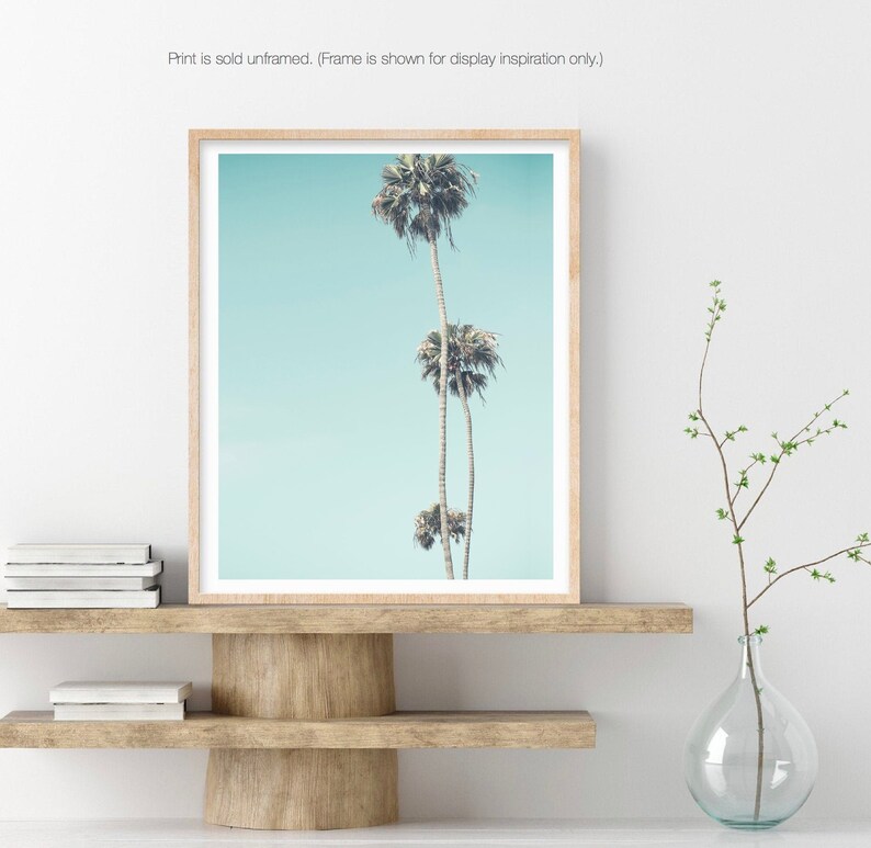 May include: A framed print of three palm trees against a light blue sky. The print is in a light wood frame and is displayed on a wooden shelf.