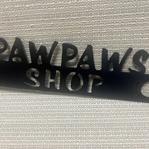 May include: Black metal sign with the words "Paw Paws Shop" cut out. The sign has two holes on either side.