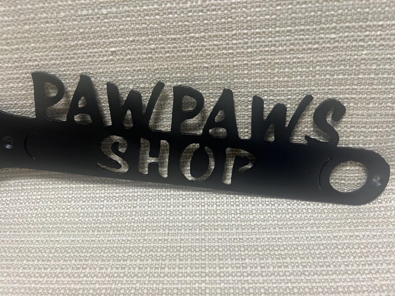 May include: Black metal sign with the words "Paw Paws Shop" cut out. The sign has two holes on either side.
