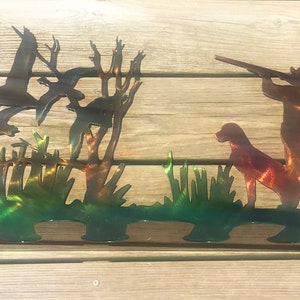 May include: Metal wall art depicting a hunter with a shotgun and a dog, standing in a marsh with ducks flying overhead. The art is painted in shades of green, brown, and orange.