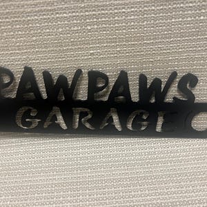 May include: Black metal sign with the words "Paw Paws Garage" cut out. The sign has two holes for hanging.