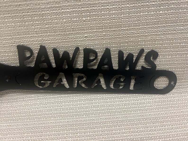 May include: Black metal sign with the words "Paw Paws Garage" cut out. The sign has two holes for hanging.