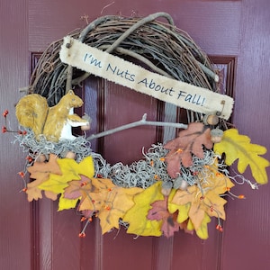 May include: A fall-themed wreath with a burlap sign that reads "I'm Nuts About Fall!" The wreath is made of twigs and has a squirrel figurine, fall leaves, and acorns.