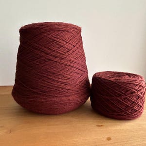 May include: Two spools of burgundy yarn. One is a large cone shape, and the other is a smaller, rounded shape. The yarn appears to be tightly wound, with visible strands creating a textured surface. Ideal for knitting and crochet projects.
