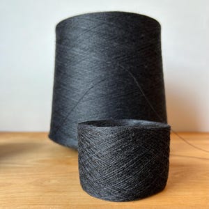 May include: Two spools of black yarn, one larger than the other, are sitting on a wooden surface. The yarn is wound tightly around the spools.