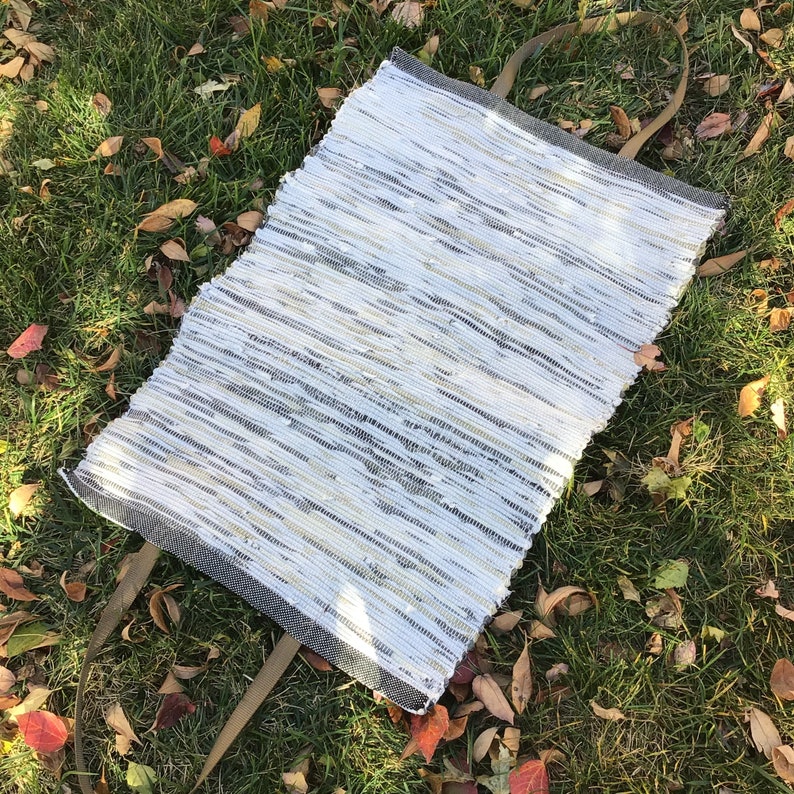 Puede incluir: Un bolso tote rectangular tejido con un patr&oacute;n de rayas blancas, grises y beige. El bolso tiene dos correas largas de color verde oliva. El bolso est&aacute; sobre un lecho de hierba verde y hojas ca&iacute;das.