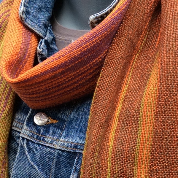 Handwoven scarf with a warp of variegated sock yarn ranging from muted green to orange to purple and a weft of orange tencel yarn.