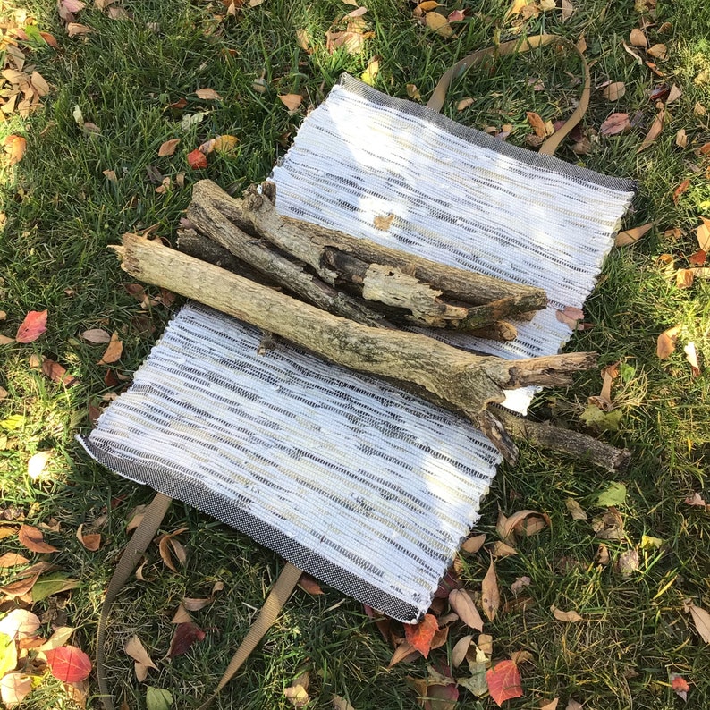 Puede incluir: Un bolso tote tejido blanco y gris con asas marrones, sobre hierba verde con hojas ca&iacute;das. Varios trozos de madera desgastada se colocan encima del bolso, sugiriendo un tema r&uacute;stico o natural.