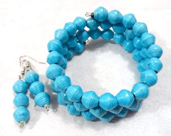 Puede incluir: Un conjunto de pulsera y pendientes de cuentas turquesas. La pulsera es de estilo envolvente, y los pendientes tienen tres cuentas cada uno. Las cuentas son de color azul claro y tienen un aspecto texturizado. El conjunto de joyas se muestra sobre una superficie blanca.