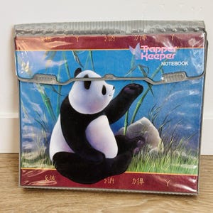 May include: A Trapper Keeper notebook featuring a graphic of a black and white panda sitting in a bamboo forest. The cover has a clear plastic overlay, a top flap with a gray plastic closure, and maroon borders with stylized characters printed on them.