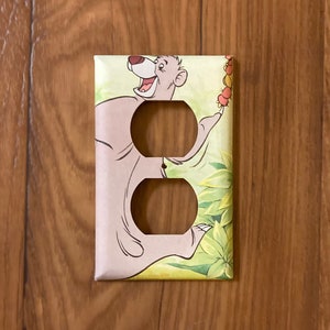 May include: A light brown and green outlet cover with a cartoon image of Baloo the bear from Disney's Jungle Book. Baloo is holding a bunch of bananas and is smiling.