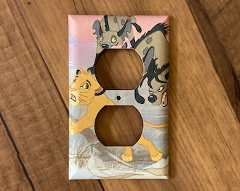 Può includere: Una placca per interruttore della luce rosa chiaro e bianco con un'immagine di cartone animato di Simba de Il Re Leone che scappa dalle iene. Le iene sono marroni e grigie con macchie nere.