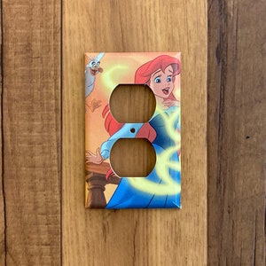 May include: A light switch cover featuring Ariel from Disney's The Little Mermaid. The cover is decorated with a colorful illustration of Ariel, a seagull, and swirling yellow light.