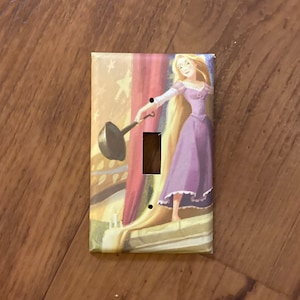 May include: A light switch cover featuring a colorful illustration of Rapunzel from the Disney movie Tangled. She is holding a frying pan and her long blonde hair is flowing down behind her.
