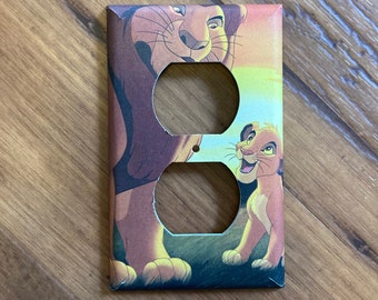 May include: A light switch cover featuring a colorful image of Simba and Mufasa from the Disney movie The Lion King. The cover has two standard outlet openings.