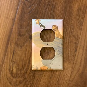 May include: Decorative electrical outlet cover featuring a scene from The Lion King. The cover has two outlet holes and depicts Simba, Rafiki, and Mufasa on a colorful background. The design is printed on a light-colored cover.