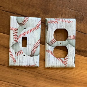 May include: Two white light switch and outlet covers with a baseball pattern. The covers have a wood grain texture and feature red baseball stitching.