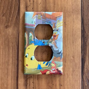 Puede incluir: Cubierta decorativa para interruptor de luz con personajes de la pel&iacute;cula animada "La Sirenita". La cubierta tiene dos recortes circulares y representa a Flounder, Sebasti&aacute;n y Scuttle en una colorida escena submarina.