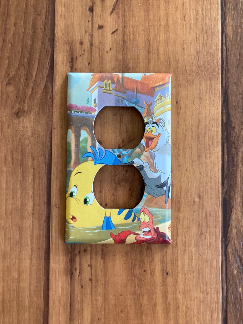 Puede incluir: Cubierta decorativa para interruptor de luz con personajes de la pel&iacute;cula animada "La Sirenita". La cubierta tiene dos recortes circulares y representa a Flounder, Sebasti&aacute;n y Scuttle en una colorida escena submarina.