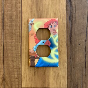 May include: A light switch cover featuring a colorful illustration of Ariel from The Little Mermaid. She is wearing a blue dress and has red hair. A seagull is flying behind her.